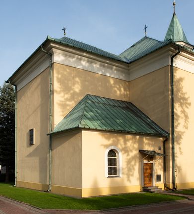 Zdjęcie nr 30: Kościół murowany, trójnawowy, halowy, na rzucie prostokąta z wydłużonym, skierowanym na północ prezbiterium z dobudowaną po zachodniej stronie zakrystią. Prezbiterium jednoprzęsłowe, węższe od korpusu, zamknięte prosto. Nawa główna oraz boczne dwuprzęsłowe, rozdzielone masywnymi opilastrowanymi filarami; artykulacja ścian przy pomocy filarów. Sklepienie prezbiterium kolebkowe, naw żagielkowe, przedzielone gurtami. Od południa chór muzyczny mieszczący się między pierwszymi filarami i powyżej kruchty wejściowej. W prezbiterium jedno okno od wschodu, drugie w zakrystii, w nawach bocznych po dwa. Główne wejście od południa flankowane przez dwa boczne, kolejne do zakrystii i między zakrystią i prezbiterium. Niektóre okna wypełnione gomułkami, ściany i sklepienia kościoła pokrywa polichromia utworzona z dekoracji floralnych i umieszczonych w jej obrębie symboli ewangelistów, herbów i putt wykonujących prace kojarzące się z regionem (bartnicze, ślusarskie, kowalskie itd.). Fasada dwuwieżowa, trójosiowa, dwuprzęsłowa, z portalem na każdej osi. Podziały pionowe przeprowadzono za pomocą półkolumienek i lizen, poziome w formie fryzu kostkowego wyniesionego nad środkowym portalem. Drzwi osi głównej w kształcie stojącego prostokąta ujętego półkolumnami dźwigającymi archiwoltę utworzoną z trzech łuków i trójkątny przyczółek. Drzwi osi bocznych zwieńczone przyczółkami. W drugiej kondygnacji na osi okulus i półokrągła konchowa wnęka z figurą Najświętszego Serca Jezusa poniżej trójkątnego szczytu dekorowanego fryzem arkadkowym. W osiach bocznych okna w kształcie stojącego prostokąta zamkniętego półkoliście. Wyżej dwie kondygnacje wież oddzielone gzymsem, w pierwszej  umieszczono okna, w drugiej tarcze zegarów. Elewacje boczne gładkie, przeprute jedynie oknami. Nawy przykrywa dach dwuspadowy, wieże namiotowe z fartuchem zwieńczone krzyżami.