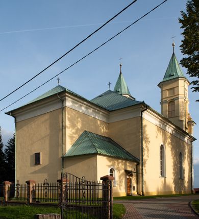 Zdjęcie nr 27: Kościół murowany, trójnawowy, halowy, na rzucie prostokąta z wydłużonym, skierowanym na północ prezbiterium z dobudowaną po zachodniej stronie zakrystią. Prezbiterium jednoprzęsłowe, węższe od korpusu, zamknięte prosto. Nawa główna oraz boczne dwuprzęsłowe, rozdzielone masywnymi opilastrowanymi filarami; artykulacja ścian przy pomocy filarów. Sklepienie prezbiterium kolebkowe, naw żagielkowe, przedzielone gurtami. Od południa chór muzyczny mieszczący się między pierwszymi filarami i powyżej kruchty wejściowej. W prezbiterium jedno okno od wschodu, drugie w zakrystii, w nawach bocznych po dwa. Główne wejście od południa flankowane przez dwa boczne, kolejne do zakrystii i między zakrystią i prezbiterium. Niektóre okna wypełnione gomułkami, ściany i sklepienia kościoła pokrywa polichromia utworzona z dekoracji floralnych i umieszczonych w jej obrębie symboli ewangelistów, herbów i putt wykonujących prace kojarzące się z regionem (bartnicze, ślusarskie, kowalskie itd.). Fasada dwuwieżowa, trójosiowa, dwuprzęsłowa, z portalem na każdej osi. Podziały pionowe przeprowadzono za pomocą półkolumienek i lizen, poziome w formie fryzu kostkowego wyniesionego nad środkowym portalem. Drzwi osi głównej w kształcie stojącego prostokąta ujętego półkolumnami dźwigającymi archiwoltę utworzoną z trzech łuków i trójkątny przyczółek. Drzwi osi bocznych zwieńczone przyczółkami. W drugiej kondygnacji na osi okulus i półokrągła konchowa wnęka z figurą Najświętszego Serca Jezusa poniżej trójkątnego szczytu dekorowanego fryzem arkadkowym. W osiach bocznych okna w kształcie stojącego prostokąta zamkniętego półkoliście. Wyżej dwie kondygnacje wież oddzielone gzymsem, w pierwszej  umieszczono okna, w drugiej tarcze zegarów. Elewacje boczne gładkie, przeprute jedynie oknami. Nawy przykrywa dach dwuspadowy, wieże namiotowe z fartuchem zwieńczone krzyżami.