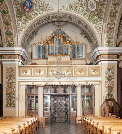 Zdjęcie nr 36: Kościół murowany, trójnawowy, halowy, na rzucie prostokąta z wydłużonym, skierowanym na północ prezbiterium z dobudowaną po zachodniej stronie zakrystią. Prezbiterium jednoprzęsłowe, węższe od korpusu, zamknięte prosto. Nawa główna oraz boczne dwuprzęsłowe, rozdzielone masywnymi opilastrowanymi filarami; artykulacja ścian przy pomocy filarów. Sklepienie prezbiterium kolebkowe, naw żagielkowe, przedzielone gurtami. Od południa chór muzyczny mieszczący się między pierwszymi filarami i powyżej kruchty wejściowej. W prezbiterium jedno okno od wschodu, drugie w zakrystii, w nawach bocznych po dwa. Główne wejście od południa flankowane przez dwa boczne, kolejne do zakrystii i między zakrystią i prezbiterium. Niektóre okna wypełnione gomułkami, ściany i sklepienia kościoła pokrywa polichromia utworzona z dekoracji floralnych i umieszczonych w jej obrębie symboli ewangelistów, herbów i putt wykonujących prace kojarzące się z regionem (bartnicze, ślusarskie, kowalskie itd.). Fasada dwuwieżowa, trójosiowa, dwuprzęsłowa, z portalem na każdej osi. Podziały pionowe przeprowadzono za pomocą półkolumienek i lizen, poziome w formie fryzu kostkowego wyniesionego nad środkowym portalem. Drzwi osi głównej w kształcie stojącego prostokąta ujętego półkolumnami dźwigającymi archiwoltę utworzoną z trzech łuków i trójkątny przyczółek. Drzwi osi bocznych zwieńczone przyczółkami. W drugiej kondygnacji na osi okulus i półokrągła konchowa wnęka z figurą Najświętszego Serca Jezusa poniżej trójkątnego szczytu dekorowanego fryzem arkadkowym. W osiach bocznych okna w kształcie stojącego prostokąta zamkniętego półkoliście. Wyżej dwie kondygnacje wież oddzielone gzymsem, w pierwszej  umieszczono okna, w drugiej tarcze zegarów. Elewacje boczne gładkie, przeprute jedynie oknami. Nawy przykrywa dach dwuspadowy, wieże namiotowe z fartuchem zwieńczone krzyżami.