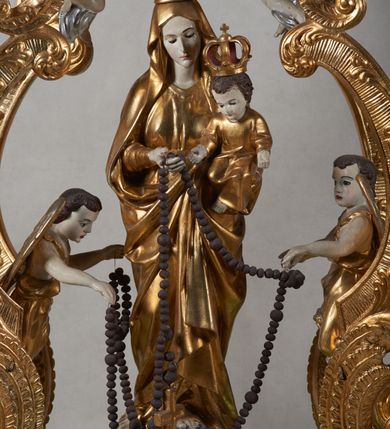 Zdjęcie nr 1: Na profilowanym, prostopadłościennym postumencie ustawiona pełnoplastyczna figura Matki Boskiej z Dzieciątkiem stojącej na niebieskim globie ziemskim, depczącej węża z czerwonym jabłkiem w pysku i podtrzymującej  różaniec. Maria jest ubrana w długie, złocone szaty, na lewym ręku podtrzymuje analogicznie ubrane Dzieciątko. Obie postaci w koronach zamkniętych. Po bokach i na górze pełnoplastyczne wizerunki aniołków siedzących na obłokach i podtrzymujących części różańca oraz różany wieniec nad głową Marii. Całość otoczona ażurową strukturą utworzoną z esowato akantowo-rocaille&#039;owych wygiętych wolut, na których ustawiony jest bogato dekorowany lambrekinem baldachim. W zwieńczeniu gloria promienista z monogramem maryjnym w obłokach. Odsłonięte partie ciała postaci polichromowane naturalistycznie, pozostałe fragmenty srebrzone i złocone; postument w kolorze jasnozielonym ze złoconymi profilami.