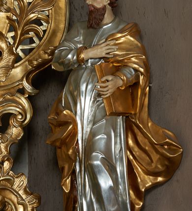 Zdjęcie nr 2: Rzeźba pełna, ustawiona frontalnie, o esowatym układzie ciała. Prawa ręka świętego spoczywa na piersi, przytrzymując płaszcz, lewa ugięta, trzyma księgę na lewym biodrze. Głowę ma zwróconą w trzech czwartych w prawo, twarz podłużną, mały nos, wyraźnie otwarte oczy i delikatnie rozchylone usta; okoloną zarostem w postaci długiej brody spływającej na pierś; włosy krótkie, w formie loków. Święty jest ubrany w obficie marszczoną tunikę, ściśle przylegającą do ciała, która spływa w dół w postaci prostych, głęboko rzeźbionych fałd. Płaszcz zawieszony na lewym ramieniu otacza postać, zawijając się na prawym udzie; tkanina ekspresyjnie poruszona, silnie łamana, drapowana w gęste fałdy. Polichromia naturalistyczna w partiach ciała, tunika srebrzona, płaszcz i księga złocone.