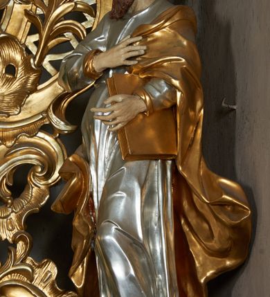 Zdjęcie nr 1: Rzeźba pełna, ustawiona frontalnie, o esowatym układzie ciała. Prawa ręka świętego spoczywa na piersi, przytrzymując płaszcz, lewa ugięta, trzyma księgę na lewym biodrze. Głowę ma zwróconą w trzech czwartych w prawo, twarz podłużną, mały nos, wyraźnie otwarte oczy i delikatnie rozchylone usta; okoloną zarostem w postaci długiej brody spływającej na pierś; włosy krótkie, w formie loków. Święty jest ubrany w obficie marszczoną tunikę, ściśle przylegającą do ciała, która spływa w dół w postaci prostych, głęboko rzeźbionych fałd. Płaszcz zawieszony na lewym ramieniu otacza postać, zawijając się na prawym udzie; tkanina ekspresyjnie poruszona, silnie łamana, drapowana w gęste fałdy. Polichromia naturalistyczna w partiach ciała, tunika srebrzona, płaszcz i księga złocone.