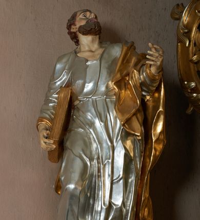 Zdjęcie nr 1: Rzeźba pełna, ustawiona frontalnie, o esowatym układzie ciała. Święty ciężar opiera na lewej nodze, prawą ma ugiętą, opartą na fragmencie skały. Lewą rękę unosi w górę, w stronę ołtarza, w wymownym geście dłoni; prawą ma opuszczoną wzdłuż boku, przytrzymuje księgę. Głowa zwrócona w trzech czwartych w lewo, uniesiona silnie w górę. Twarz podłużna o wyraźnych, głęboko rzeźbionych rysach, wąskim nosie, szeroko otwartych, dużych oczach skierowanych w górę; okolona zarostem; włosy krótkie, delikatnie falowane, opadają na tył głowy. Postać ubrana jest w tunikę przepasaną w talii; tkanina jest silnie drapowana, ściśle przylega do ciała, odzwierciedlając jego układ, opadając w dół w formie gęstych fałd. Święty ma płaszcz przewieszony przez lewe ramię, spływa na plecy w formie ciężkiej, silnie drapowanej i łamanej tkaniny. Polichromia naturalistyczna w partiach ciała, tunika srebrzona, płaszcz oraz księga złocone.