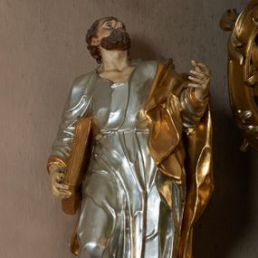 Zdjęcie nr 1: Rzeźba pełna, ustawiona frontalnie, o esowatym układzie ciała. Święty ciężar opiera na lewej nodze, prawą ma ugiętą, opartą na fragmencie skały. Lewą rękę unosi w górę, w stronę ołtarza, w wymownym geście dłoni; prawą ma opuszczoną wzdłuż boku, przytrzymuje księgę. Głowa zwrócona w trzech czwartych w lewo, uniesiona silnie w górę. Twarz podłużna o wyraźnych, głęboko rzeźbionych rysach, wąskim nosie, szeroko otwartych, dużych oczach skierowanych w górę; okolona zarostem; włosy krótkie, delikatnie falowane, opadają na tył głowy. Postać ubrana jest w tunikę przepasaną w talii; tkanina jest silnie drapowana, ściśle przylega do ciała, odzwierciedlając jego układ, opadając w dół w formie gęstych fałd. Święty ma płaszcz przewieszony przez lewe ramię, spływa na plecy w formie ciężkiej, silnie drapowanej i łamanej tkaniny. Polichromia naturalistyczna w partiach ciała, tunika srebrzona, płaszcz oraz księga złocone.
