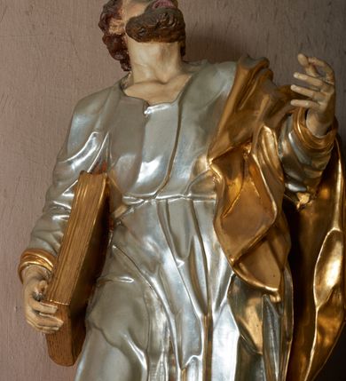 Zdjęcie nr 2: Rzeźba pełna, ustawiona frontalnie, o esowatym układzie ciała. Święty ciężar opiera na lewej nodze, prawą ma ugiętą, opartą na fragmencie skały. Lewą rękę unosi w górę, w stronę ołtarza, w wymownym geście dłoni; prawą ma opuszczoną wzdłuż boku, przytrzymuje księgę. Głowa zwrócona w trzech czwartych w lewo, uniesiona silnie w górę. Twarz podłużna o wyraźnych, głęboko rzeźbionych rysach, wąskim nosie, szeroko otwartych, dużych oczach skierowanych w górę; okolona zarostem; włosy krótkie, delikatnie falowane, opadają na tył głowy. Postać ubrana jest w tunikę przepasaną w talii; tkanina jest silnie drapowana, ściśle przylega do ciała, odzwierciedlając jego układ, opadając w dół w formie gęstych fałd. Święty ma płaszcz przewieszony przez lewe ramię, spływa na plecy w formie ciężkiej, silnie drapowanej i łamanej tkaniny. Polichromia naturalistyczna w partiach ciała, tunika srebrzona, płaszcz oraz księga złocone.