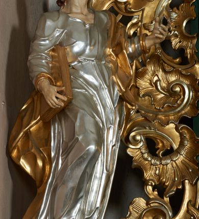 Zdjęcie nr 3: Rzeźba pełna, ustawiona frontalnie, o esowatym układzie ciała. Święty ciężar opiera na lewej nodze, prawą ma ugiętą, opartą na fragmencie skały. Lewą rękę unosi w górę, w stronę ołtarza, w wymownym geście dłoni; prawą ma opuszczoną wzdłuż boku, przytrzymuje księgę. Głowa zwrócona w trzech czwartych w lewo, uniesiona silnie w górę. Twarz podłużna o wyraźnych, głęboko rzeźbionych rysach, wąskim nosie, szeroko otwartych, dużych oczach skierowanych w górę; okolona zarostem; włosy krótkie, delikatnie falowane, opadają na tył głowy. Postać ubrana jest w tunikę przepasaną w talii; tkanina jest silnie drapowana, ściśle przylega do ciała, odzwierciedlając jego układ, opadając w dół w formie gęstych fałd. Święty ma płaszcz przewieszony przez lewe ramię, spływa na plecy w formie ciężkiej, silnie drapowanej i łamanej tkaniny. Polichromia naturalistyczna w partiach ciała, tunika srebrzona, płaszcz oraz księga złocone.