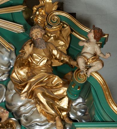 Zdjęcie nr 4: Grupa płaskorzeźbiona złożona z figury Chrystusa i Boga Ojca w otoczeniu obłoków oraz gołębicy Ducha Świętego. Po lewej stronie postać siedzącego Chrystusa, zwrócona w trzech czwartych do wewnątrz. Jego lewa ręka spoczywa na piersi, prawą przytrzymuje krzyż. Ma twarz o wyraźnych rysach, małym nosie oraz szeroko otwartych oczach; okoloną zarostem; włosy długie, opadają na plecy w postaci fal. Ciało o wyraźnie zarysowanej anatomii. Figura w partii bioder przepasana jest płaszczem spływającym na nogi i zawieszonym na prawym ramieniu; tkanina obficie drapowana w duże fałdy i delikatne załamania. Po prawej stronie siedząca postać Boga Ojca, zwrócona w trzech czwartych na zewnątrz. Lewą rękę wysuwa w bok, prawą w geście błogosławieństwa wyciąga przed siebie. Głowę ma skierowaną w prawą stronę, delikatnie pochyloną, spogląda w dół. Ma podłużną twarz o starczych rysach i niewielkim nosie, która jest okolona długą siwą brodą, spływającą na pierś; włosy krótkie, delikatnie falowane. Postać ubrana w tunikę, przepasaną w talii, o obficie marszczonej tkaninie, ściśle przylegającej do ciała; płaszcz zawinięty na prawym ramieniu, spoczywa na kolanach w formie obficie fałdowanej, ciężkiej draperii. Między postaciami kula w otoczeniu obłoków i uskrzydlonych główek anielskich. Całość wieńczy gołębica Ducha Świętego w glorii promienistej. Polichromia naturalistyczna w partiach ciała; obłoki, kula i gołębica srebrzone; szaty, atrybuty i gloria złocone.