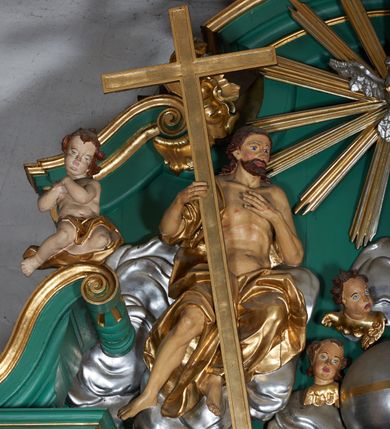 Zdjęcie nr 5: Grupa płaskorzeźbiona złożona z figury Chrystusa i Boga Ojca w otoczeniu obłoków oraz gołębicy Ducha Świętego. Po lewej stronie postać siedzącego Chrystusa, zwrócona w trzech czwartych do wewnątrz. Jego lewa ręka spoczywa na piersi, prawą przytrzymuje krzyż. Ma twarz o wyraźnych rysach, małym nosie oraz szeroko otwartych oczach; okoloną zarostem; włosy długie, opadają na plecy w postaci fal. Ciało o wyraźnie zarysowanej anatomii. Figura w partii bioder przepasana jest płaszczem spływającym na nogi i zawieszonym na prawym ramieniu; tkanina obficie drapowana w duże fałdy i delikatne załamania. Po prawej stronie siedząca postać Boga Ojca, zwrócona w trzech czwartych na zewnątrz. Lewą rękę wysuwa w bok, prawą w geście błogosławieństwa wyciąga przed siebie. Głowę ma skierowaną w prawą stronę, delikatnie pochyloną, spogląda w dół. Ma podłużną twarz o starczych rysach i niewielkim nosie, która jest okolona długą siwą brodą, spływającą na pierś; włosy krótkie, delikatnie falowane. Postać ubrana w tunikę, przepasaną w talii, o obficie marszczonej tkaninie, ściśle przylegającej do ciała; płaszcz zawinięty na prawym ramieniu, spoczywa na kolanach w formie obficie fałdowanej, ciężkiej draperii. Między postaciami kula w otoczeniu obłoków i uskrzydlonych główek anielskich. Całość wieńczy gołębica Ducha Świętego w glorii promienistej. Polichromia naturalistyczna w partiach ciała; obłoki, kula i gołębica srebrzone; szaty, atrybuty i gloria złocone.
