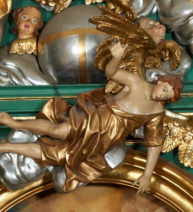 Zdjęcie nr 6: Grupa płaskorzeźbiona złożona z figury Chrystusa i Boga Ojca w otoczeniu obłoków oraz gołębicy Ducha Świętego. Po lewej stronie postać siedzącego Chrystusa, zwrócona w trzech czwartych do wewnątrz. Jego lewa ręka spoczywa na piersi, prawą przytrzymuje krzyż. Ma twarz o wyraźnych rysach, małym nosie oraz szeroko otwartych oczach; okoloną zarostem; włosy długie, opadają na plecy w postaci fal. Ciało o wyraźnie zarysowanej anatomii. Figura w partii bioder przepasana jest płaszczem spływającym na nogi i zawieszonym na prawym ramieniu; tkanina obficie drapowana w duże fałdy i delikatne załamania. Po prawej stronie siedząca postać Boga Ojca, zwrócona w trzech czwartych na zewnątrz. Lewą rękę wysuwa w bok, prawą w geście błogosławieństwa wyciąga przed siebie. Głowę ma skierowaną w prawą stronę, delikatnie pochyloną, spogląda w dół. Ma podłużną twarz o starczych rysach i niewielkim nosie, która jest okolona długą siwą brodą, spływającą na pierś; włosy krótkie, delikatnie falowane. Postać ubrana w tunikę, przepasaną w talii, o obficie marszczonej tkaninie, ściśle przylegającej do ciała; płaszcz zawinięty na prawym ramieniu, spoczywa na kolanach w formie obficie fałdowanej, ciężkiej draperii. Między postaciami kula w otoczeniu obłoków i uskrzydlonych główek anielskich. Całość wieńczy gołębica Ducha Świętego w glorii promienistej. Polichromia naturalistyczna w partiach ciała; obłoki, kula i gołębica srebrzone; szaty, atrybuty i gloria złocone.