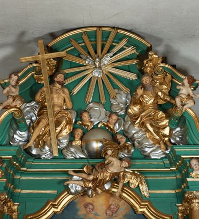 Zdjęcie nr 1: Grupa płaskorzeźbiona złożona z figury Chrystusa i Boga Ojca w otoczeniu obłoków oraz gołębicy Ducha Świętego. Po lewej stronie postać siedzącego Chrystusa, zwrócona w trzech czwartych do wewnątrz. Jego lewa ręka spoczywa na piersi, prawą przytrzymuje krzyż. Ma twarz o wyraźnych rysach, małym nosie oraz szeroko otwartych oczach; okoloną zarostem; włosy długie, opadają na plecy w postaci fal. Ciało o wyraźnie zarysowanej anatomii. Figura w partii bioder przepasana jest płaszczem spływającym na nogi i zawieszonym na prawym ramieniu; tkanina obficie drapowana w duże fałdy i delikatne załamania. Po prawej stronie siedząca postać Boga Ojca, zwrócona w trzech czwartych na zewnątrz. Lewą rękę wysuwa w bok, prawą w geście błogosławieństwa wyciąga przed siebie. Głowę ma skierowaną w prawą stronę, delikatnie pochyloną, spogląda w dół. Ma podłużną twarz o starczych rysach i niewielkim nosie, która jest okolona długą siwą brodą, spływającą na pierś; włosy krótkie, delikatnie falowane. Postać ubrana w tunikę, przepasaną w talii, o obficie marszczonej tkaninie, ściśle przylegającej do ciała; płaszcz zawinięty na prawym ramieniu, spoczywa na kolanach w formie obficie fałdowanej, ciężkiej draperii. Między postaciami kula w otoczeniu obłoków i uskrzydlonych główek anielskich. Całość wieńczy gołębica Ducha Świętego w glorii promienistej. Polichromia naturalistyczna w partiach ciała; obłoki, kula i gołębica srebrzone; szaty, atrybuty i gloria złocone.