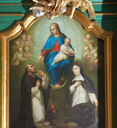Zdjęcie nr 5: Obraz w formie stojącego prostokąta zamkniętego łukiem nadwieszonym z uskokiem. W centrum tronuje Maria z Dzieciątkiem na kolanach i półksiężycem pod prawą stopą. Postać jest ustawiona frontalnie, delikatnie pochyla głowę w prawo. Prawą ręką wysuniętą w przód podaje różaniec św. Dominikowi, lewą obejmuje Dzieciątko. Maria ma twarz o młodzieńczych, delikatnych rysach, z wyraźnie zarysowanym długim nosem, półprzymkniętymi powiekami. Jasne włosy z przedziałkiem pośrodku, opadają  zwinięte w pukle na ramiona i plecy. Maria jest ubrana w czerwoną tunikę przepasaną w talii oraz niebieski płaszcz spięty na piersi zaponą, przerzucony z przodu przez kolana. Dzieciątko zwrócone w trzech czwartych w prawo, z korpusem skierowanym w lewo; delikatnie pochylone; obiema dłońmi trzyma wieniec z kwiatów nad głową św. Katarzyny. Ma okrągłą twarz o wyraźnych rysach, wysokie czoło, okrągłe oczy, małe usta i nos; włosy krótkie, jasne, układające się w loki. Jest ubrane w białą tunikę w podwiniętymi rękawami. Po lewej stronie, klęcząca postać św. Dominika, ukazanego w całej postaci, z prawego profilu. Święty prawą rękę składa na piersi, lewą odbiera różaniec od Marii. Głowę ma uniesioną lekko ku górze, twarz szczupłą, o ostrym profilu nosa, wzrok kieruje na Marię; włosy krótkie z tonsurą, krótka broda. Święty ubrany jest w habit dominikański. U jego stóp brązowy pies z pochodnią w pysku. Po prawej stronie obrazu postać klęczącej św. Katarzyny Sieneńskiej ukazanej w całej postaci, zwróconej w prawo, z lewą ręką opuszczoną, prawą zaś złożoną na piersi. Twarz świętej jest owalna, o dziewczęcych rysach i jasnej karnacji, wzrok skierowany ku górze w stronę Marii, nos długi o prostym profilu, usta niewielkie. U góry kompozycji, po bokach Marii uskrzydlone, nagie putta trzymające różane wianki. Tło szaro-zielonkawe wypełnione powtarzającym się motywem główek anielskich.