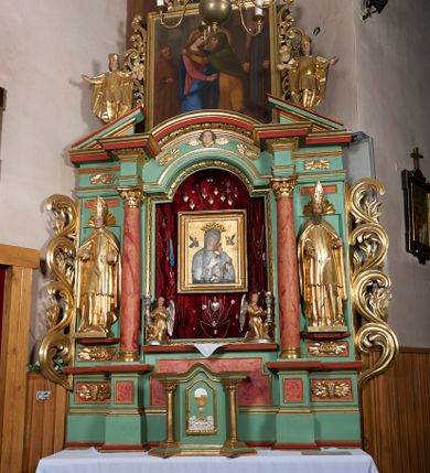 Zdjęcie nr 1: Ołtarz przyścienny, architektoniczny, trójosiowy, jednokondygnacyjny ze zwieńczeniem i uszakami. Mensa w typie sarkofagowym na niewielkim prostopadłościennym postumencie. Tabernakulum drewniane, architektoniczne, ujęte złoconymi kolumienkami dźwigającymi wyłamane i wyniesione belkowanie. Na drzwiczkach płaskorzeźbione przedstawienie kielicha z hostią. Nastawa drewniana ujęta korynckimi kolumnami ustawionymi na wysokich postumentach. Dźwigają one pełne, wyłamane na osiach kolumn i półkoliście wyniesione na osi środkowej ołtarza belkowanie. Fryz dekorowany rozetami oraz uskrzydloną główką anielską. Powyżej odcinki przyczółka, na których ustawione pełnoplastyczne figury świętych wojowników. W polu środkowym nisza zamknięta łukiem półkolistym nadwieszonym mieszcząca obraz Matki Boskiej Nieustającej Pomocy. W osiach bocznych ustawione na tle płycin pełnoplastyczne figury świętych biskupów w złoconych szatach oraz akantowe, ażurowe uszaki utworzone z wici akantowej. W zwieńczeniu obraz „Nawiedzenie Najświętszej Marii Panny” w kształcie stojącego prostokąta ujęty ażurowymi uszami utworzonymi z ornamentu mażowinowo-chrząstkowego, i winorośli. Powyżej, analogicznie, mniejszy obraz „Trójca Święta” w kształcie prostokąta stojącego o ściętych górnych narożach, zwieńczonego krzyżem równoramiennym. Struktura ołtarza w kolorze jasnozielonym, wypełnienia płycin, trzony kolumn oraz elementy gzymsu jasnoczerwone, marmoryzowane; ornamenty złocone.