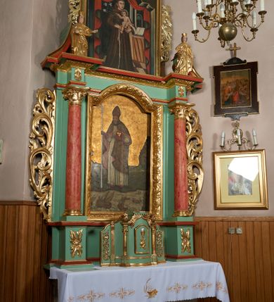 Zdjęcie nr 3: Ołtarz przyścienny, architektoniczny, jednoosiowy, jednokondygnacyjny ze zwieńczeniem; uszaty. Mensa w typie sarkofagowym na niewielkim prostopadłościennym postumencie. Tabernakulum drewniane, architektoniczne, ujęte parami złoconych kolumienek, na osi których odcinki belkowania i spiralnie zwinięte akantowe woluty oraz kanelowanymi pilastrami. W zwieńczeniu motyw podwieszonej kotary oraz lambrekin. Na drzwiczkach płaskorzeźbione przedstawienie kielicha z hostią. Nastawa drewniana, ujęta korynckimi kolumnami ustawionymi na wysokich postumentach zdobionych plakietami roślinnymi. Dźwigają one pełne, wyłamane na ich osiach belkowanie. Fryz wysuniętych części dekorowany rozetami, silnie wystający gzyms zdobiony akantem. Powyżej odcinki przyczółka, na tle których ustawione pełnoplastyczne figury świętych dziewic w koronach i złoconych szatach. W kondygnacji pierwszej nisza zamknięta łukiem półkolistym nadwieszonym, mieszcząca obraz św. Mikołaja. Kondygnacja ujęta ażurowymi akantowymi uszakami. W zwieńczeniu przedstawienie św. Antoniego Padewskiego w formie stojącego prostokąta, ujętego ażurowymi uszakami utworzonymi z wici akantu. Powyżej, analogicznie, mniejsze ujęcie Boga Ojca w kształcie stojącego prostokąta o ściętych górnych narożach, zwieńczonego krzyżem równoramiennym. Struktura ołtarza w kolorze jasnozielonym, wypełnienia płycin, trzony kolumn oraz elementy gzymsu jasnoczerwone; ornamenty złocone.