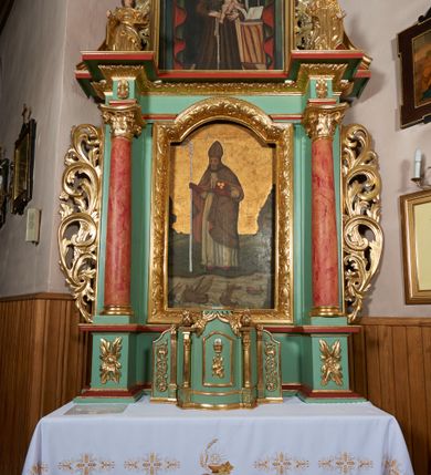 Zdjęcie nr 4: Ołtarz przyścienny, architektoniczny, jednoosiowy, jednokondygnacyjny ze zwieńczeniem; uszaty. Mensa w typie sarkofagowym na niewielkim prostopadłościennym postumencie. Tabernakulum drewniane, architektoniczne, ujęte parami złoconych kolumienek, na osi których odcinki belkowania i spiralnie zwinięte akantowe woluty oraz kanelowanymi pilastrami. W zwieńczeniu motyw podwieszonej kotary oraz lambrekin. Na drzwiczkach płaskorzeźbione przedstawienie kielicha z hostią. Nastawa drewniana, ujęta korynckimi kolumnami ustawionymi na wysokich postumentach zdobionych plakietami roślinnymi. Dźwigają one pełne, wyłamane na ich osiach belkowanie. Fryz wysuniętych części dekorowany rozetami, silnie wystający gzyms zdobiony akantem. Powyżej odcinki przyczółka, na tle których ustawione pełnoplastyczne figury świętych dziewic w koronach i złoconych szatach. W kondygnacji pierwszej nisza zamknięta łukiem półkolistym nadwieszonym, mieszcząca obraz św. Mikołaja. Kondygnacja ujęta ażurowymi akantowymi uszakami. W zwieńczeniu przedstawienie św. Antoniego Padewskiego w formie stojącego prostokąta, ujętego ażurowymi uszakami utworzonymi z wici akantu. Powyżej, analogicznie, mniejsze ujęcie Boga Ojca w kształcie stojącego prostokąta o ściętych górnych narożach, zwieńczonego krzyżem równoramiennym. Struktura ołtarza w kolorze jasnozielonym, wypełnienia płycin, trzony kolumn oraz elementy gzymsu jasnoczerwone; ornamenty złocone.