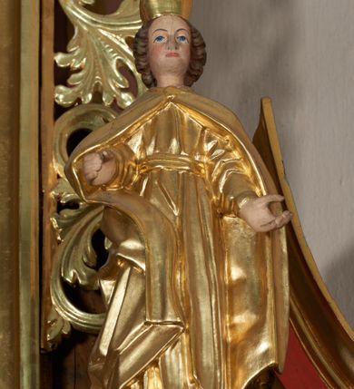 Zdjęcie nr 11: Ołtarz przyścienny, architektoniczny, jednoosiowy, jednokondygnacyjny ze zwieńczeniem; uszaty. Mensa w typie sarkofagowym na niewielkim prostopadłościennym postumencie. Tabernakulum drewniane, architektoniczne, ujęte parami złoconych kolumienek, na osi których odcinki belkowania i spiralnie zwinięte akantowe woluty oraz kanelowanymi pilastrami. W zwieńczeniu motyw podwieszonej kotary oraz lambrekin. Na drzwiczkach płaskorzeźbione przedstawienie kielicha z hostią. Nastawa drewniana, ujęta korynckimi kolumnami ustawionymi na wysokich postumentach zdobionych plakietami roślinnymi. Dźwigają one pełne, wyłamane na ich osiach belkowanie. Fryz wysuniętych części dekorowany rozetami, silnie wystający gzyms zdobiony akantem. Powyżej odcinki przyczółka, na tle których ustawione pełnoplastyczne figury świętych dziewic w koronach i złoconych szatach. W kondygnacji pierwszej nisza zamknięta łukiem półkolistym nadwieszonym, mieszcząca obraz św. Mikołaja. Kondygnacja ujęta ażurowymi akantowymi uszakami. W zwieńczeniu przedstawienie św. Antoniego Padewskiego w formie stojącego prostokąta, ujętego ażurowymi uszakami utworzonymi z wici akantu. Powyżej, analogicznie, mniejsze ujęcie Boga Ojca w kształcie stojącego prostokąta o ściętych górnych narożach, zwieńczonego krzyżem równoramiennym. Struktura ołtarza w kolorze jasnozielonym, wypełnienia płycin, trzony kolumn oraz elementy gzymsu jasnoczerwone; ornamenty złocone.