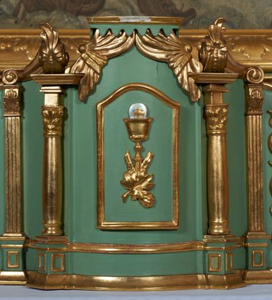 Zdjęcie nr 12: Ołtarz przyścienny, architektoniczny, jednoosiowy, jednokondygnacyjny ze zwieńczeniem; uszaty. Mensa w typie sarkofagowym na niewielkim prostopadłościennym postumencie. Tabernakulum drewniane, architektoniczne, ujęte parami złoconych kolumienek, na osi których odcinki belkowania i spiralnie zwinięte akantowe woluty oraz kanelowanymi pilastrami. W zwieńczeniu motyw podwieszonej kotary oraz lambrekin. Na drzwiczkach płaskorzeźbione przedstawienie kielicha z hostią. Nastawa drewniana, ujęta korynckimi kolumnami ustawionymi na wysokich postumentach zdobionych plakietami roślinnymi. Dźwigają one pełne, wyłamane na ich osiach belkowanie. Fryz wysuniętych części dekorowany rozetami, silnie wystający gzyms zdobiony akantem. Powyżej odcinki przyczółka, na tle których ustawione pełnoplastyczne figury świętych dziewic w koronach i złoconych szatach. W kondygnacji pierwszej nisza zamknięta łukiem półkolistym nadwieszonym, mieszcząca obraz św. Mikołaja. Kondygnacja ujęta ażurowymi akantowymi uszakami. W zwieńczeniu przedstawienie św. Antoniego Padewskiego w formie stojącego prostokąta, ujętego ażurowymi uszakami utworzonymi z wici akantu. Powyżej, analogicznie, mniejsze ujęcie Boga Ojca w kształcie stojącego prostokąta o ściętych górnych narożach, zwieńczonego krzyżem równoramiennym. Struktura ołtarza w kolorze jasnozielonym, wypełnienia płycin, trzony kolumn oraz elementy gzymsu jasnoczerwone; ornamenty złocone.