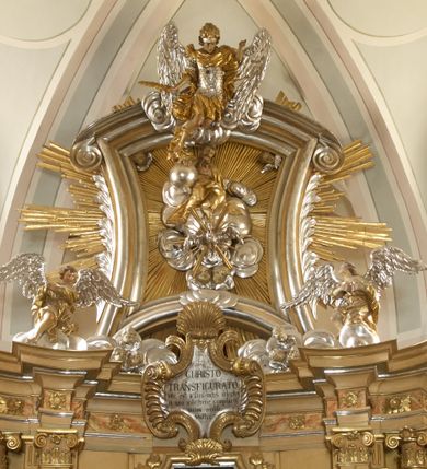 Zdjęcie nr 5: Ołtarz architektoniczny, jednokondygnacyjny, jednoosiowy, ze zwieńczeniem. Nastawa zbudowana na kamiennym, wysokim cokole, lekko wklęsła, ujęta zwielokrotnionymi pilastrami kompozytowymi o kanelowanych trzonach i zewnętrznymi, nieznacznie cofniętymi kolumnami, podtrzymującymi wyłamujące się nad nimi belkowanie. Po bokach karbowane taśmy z motywem liści akantu. W centrum obraz "Przemienienie Pańskie". Nad pilastrami klęczące na obłokach uskrzydlone, adorujące anioły. Zwieńczenie o łukowato wygiętej podstawie i wklęsłych nieznacznie bokach, zwieńczone wyłamanym ku górze gzymsem zakończonym po bokach wolutami. W zwieńczenie wkomponowana okazała gloria promienista, z figurą Boga Ojca siedzącego w chmurach, Gołębicą Ducha Świętego i ukazaną na tle gzymsu postacią Archanioła Michała, ukazanego w zbroi, płaszczu na ramionach i w hełmie na głowie z mieczem w prawej dłoni. Na belkowaniu okazały kartusz ujęty puklowaną wstęgą i zwieńczony muszlą, z malowanym na srebrnym tle napisem: "CHRISTO / TRANSFIGURATO / Hic est Filius meus dilectus / in quo mihi bene complacui / ipsum audite. / Matth: c. 77". Na ścianach prezbiterium, przed ołtarzem, na konsolach dwie figury św. Andrzej i św. Mateusz (?). Nastawa w całości złocona, partiami srebrzona (chmury, skrzydła aniołów, rama zwieńczenia, wstęga regencyjna po bokach), partie cokołu i fryzu z imitacją marmoryzacji.   

