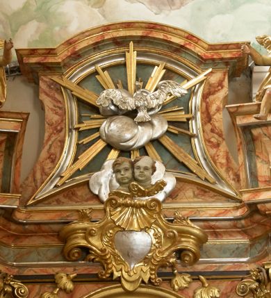 Zdjęcie nr 15: Retabulum architektoniczne, jednoosiowe, jednokondygnacyjne, ze zwieńczeniem, wzniesione na wysokim cokole. Nastawa ujęta ukośnie ustawionymi połówkami filarów na tle pilastrów, o kapitelach kompozytowych, podtrzymujących wydatne, przełamujące się nad nimi belkowanie z impostami i zwróconymi ku przodowi naczółkami, na których siedzą uskrzydlone, nagie putta, przepasane tkaninami. Zwieńczenie o lekko wklęsłych bokach zakończone gzymsem w formie łuku nadwieszonego. W partii fryzu kartusz ujęty wolutami, z lambrekinem u dołu z muszlą palmetową u góry. W zwieńczeniu gołębica Ducha Świętego na tle obłoków i promienistej glorii. Dekorację ornamentalną tworzą kratki regencyjne, kampanule i muszle palmetowe. W części środkowej obraz Matki Boskiej z Dzieciątkiem w typie Matki Boskiej Śnieżnej w drewnianej, złoconej sukience z koronami na głowie. Obraz z profilowanej ramie, złoconej i srebrzonej, ujętej po bokach zwisami z liści i kwiatów. Ponad obrazem rozłożysta muszla palmetowa. Po bokach nastawy dwie niezidentyfikowane postacie mężczyzn, całopostaciowe, w długich sukniach, z płaszczami zarzuconymi na ramiona, jedna z otwartą księgą w prawej dłoni, druga z rękoma złożonymi w geście modlitwy. Polichromia imitująca marmoryzację w odcieniach jasnych brązów, czerwieni i zieleni, figury o naturalnych karnacjach, złocenia szat figur i ornamentów, srebrzenia drobnych detali ornamentalnych.      


