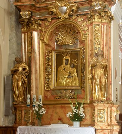 Zdjęcie nr 2: Retabulum architektoniczne, jednoosiowe, jednokondygnacyjne, ze zwieńczeniem, wzniesione na wysokim cokole. Nastawa ujęta ukośnie ustawionymi połówkami filarów na tle pilastrów, o kapitelach kompozytowych, podtrzymujących wydatne, przełamujące się nad nimi belkowanie z impostami i zwróconymi ku przodowi naczółkami, na których siedzą uskrzydlone, nagie putta, przepasane tkaninami. Zwieńczenie o lekko wklęsłych bokach zakończone gzymsem w formie łuku nadwieszonego. W partii fryzu kartusz ujęty wolutami, z lambrekinem u dołu z muszlą palmetową u góry. W zwieńczeniu gołębica Ducha Świętego na tle obłoków i promienistej glorii. Dekorację ornamentalną tworzą kratki regencyjne, kampanule i muszle palmetowe. W części środkowej obraz Matki Boskiej z Dzieciątkiem w typie Matki Boskiej Śnieżnej w drewnianej, złoconej sukience z koronami na głowie. Obraz z profilowanej ramie, złoconej i srebrzonej, ujętej po bokach zwisami z liści i kwiatów. Ponad obrazem rozłożysta muszla palmetowa. Po bokach nastawy dwie niezidentyfikowane postacie mężczyzn, całopostaciowe, w długich sukniach, z płaszczami zarzuconymi na ramiona, jedna z otwartą księgą w prawej dłoni, druga z rękoma złożonymi w geście modlitwy. Polichromia imitująca marmoryzację w odcieniach jasnych brązów, czerwieni i zieleni, figury o naturalnych karnacjach, złocenia szat figur i ornamentów, srebrzenia drobnych detali ornamentalnych.      


