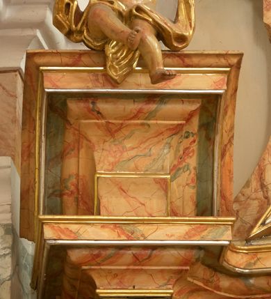 Zdjęcie nr 19: Retabulum architektoniczne, jednoosiowe, jednokondygnacyjne, ze zwieńczeniem, wzniesione na wysokim cokole. Nastawa ujęta ukośnie ustawionymi połówkami filarów na tle pilastrów, o kapitelach kompozytowych, podtrzymujących wydatne, przełamujące się nad nimi belkowanie z impostami i zwróconymi ku przodowi naczółkami, na których siedzą uskrzydlone, nagie putta, przepasane tkaninami. Zwieńczenie o lekko wklęsłych bokach zakończone gzymsem w formie łuku nadwieszonego. W partii fryzu kartusz ujęty wolutami, z lambrekinem u dołu z muszlą palmetową u góry. W zwieńczeniu gołębica Ducha Świętego na tle obłoków i promienistej glorii. Dekorację ornamentalną tworzą kratki regencyjne, kampanule i muszle palmetowe. W części środkowej obraz Matki Boskiej z Dzieciątkiem w typie Matki Boskiej Śnieżnej w drewnianej, złoconej sukience z koronami na głowie. Obraz z profilowanej ramie, złoconej i srebrzonej, ujętej po bokach zwisami z liści i kwiatów. Ponad obrazem rozłożysta muszla palmetowa. Po bokach nastawy dwie niezidentyfikowane postacie mężczyzn, całopostaciowe, w długich sukniach, z płaszczami zarzuconymi na ramiona, jedna z otwartą księgą w prawej dłoni, druga z rękoma złożonymi w geście modlitwy. Polichromia imitująca marmoryzację w odcieniach jasnych brązów, czerwieni i zieleni, figury o naturalnych karnacjach, złocenia szat figur i ornamentów, srebrzenia drobnych detali ornamentalnych.      


