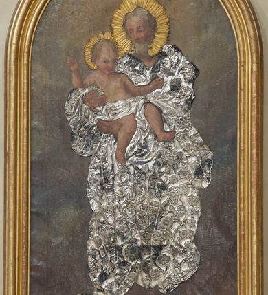 Zdjęcie nr 1: Obraz w kształcie stojącego prostokąta zamkniętego półkoliście w drewnianej, profilowanej, złoconej ramie. W centrum przedstawienia znajduje się stojący frontalnie w lekkim kontrapoście św. Józef trzymający oburącz Dzieciątko Jezus. Święty spogląda czule na Jezusa, ma owalną, łagodną twarz, migdałowe, półprzymknięte oczy, wąski nos, małe usta oraz delikatne rumieńce. Dzieciątko patrzy na widza, lewą rękę wyciąga do boku, prawą unosi w geście błogosławieństwa, zgięte w kolanach nogi ma luźno opuszczone. Na okrągłej twarzy wyróżniają się bystre, ciemne oczy, mały nos, wąskie usta oraz rumieńce. Święty Józef przyodziany jest w nałożoną na obraz metalową, długą, pofałdowaną, suknię pokrytą ornamentem roślinno-kwiatowym, która przechodzi w przepaskę na biodrach Jezusa. Głowy postaci otaczają aplikowane, złocone nimby promieniste. Scena rozgrywa się na ciemnym, rozjaśnionym wokół głów postaci tle.
