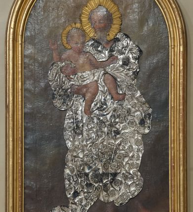 Zdjęcie nr 2: Obraz w kształcie stojącego prostokąta zamkniętego półkoliście w drewnianej, profilowanej, złoconej ramie. W centrum przedstawienia znajduje się stojący frontalnie w lekkim kontrapoście św. Józef trzymający oburącz Dzieciątko Jezus. Święty spogląda czule na Jezusa, ma owalną, łagodną twarz, migdałowe, półprzymknięte oczy, wąski nos, małe usta oraz delikatne rumieńce. Dzieciątko patrzy na widza, lewą rękę wyciąga do boku, prawą unosi w geście błogosławieństwa, zgięte w kolanach nogi ma luźno opuszczone. Na okrągłej twarzy wyróżniają się bystre, ciemne oczy, mały nos, wąskie usta oraz rumieńce. Święty Józef przyodziany jest w nałożoną na obraz metalową, długą, pofałdowaną, suknię pokrytą ornamentem roślinno-kwiatowym, która przechodzi w przepaskę na biodrach Jezusa. Głowy postaci otaczają aplikowane, złocone nimby promieniste. Scena rozgrywa się na ciemnym, rozjaśnionym wokół głów postaci tle.