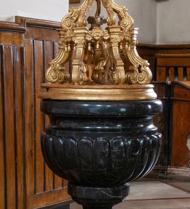 Zdjęcie nr 1: Chrzcielnica osadzona na wysokim, dwuczęściowym, schodkowym postumencie, z trzonem w kształcie gruszkowatym i puklowaną czarą z profilowanym wylewem. Pokrywa utworzona z ośmiu drewnianych kabłąków w kształcie dwóch rzędów spływów wolutowych, ozdobionych motywem koguciego grzebienia, podtrzymujących glob ziemski nakryty koroną zamkniętą z czterema kabłąkami z kulą i krzyżem. Wewnątrz ażurowej pokrywy grupa rzeźbiarska Chrzest Chrystusa w Jordanie i gołębica Ducha Świętego nad nimi.