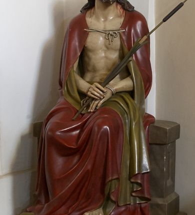 Zdjęcie nr 1: Rzeźba przedstawia Jezusa w typie Ecce Homo, po biczowaniu, ze śladami męki, siedzącego na kamieniu z rękami złożonymi na lewym udzie. Zbawiciel z bolesnym wyrazem twarzy zwraca wzrok ku górze, ma owalną twarz, duże, jasne oczy otoczone opuchniętymi powiekami spoglądające spod uniesionych brwi, wąski nos oraz niewielkie, nieznacznie rozchylone usta. Twarz okala ciemny zarost, spod korony cierniowej nałożonej na długie, opadające na ramiona ciemne włosy spływają strużki krwi. W prawej dłoni związanych sznurem rąk trzyma trzcinę, lewą stopę opiera na kamieniu, prawą na ziemi. Spod okrywającego jego plecy i nogi, związanego pod szyją purpurowego, podbitego złotem płaszcza widać nagi, umięśniony tors.