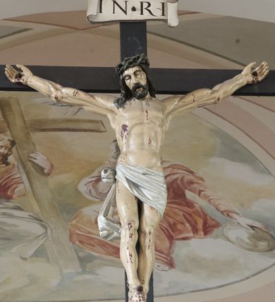 Zdjęcie nr 2: Krucyfiks to przedstawienie ukrzyżowanego Jezusa. Martwe ciało Zbawiciela o wyraźnie zaznaczonej muskulaturze jest rozciągnięte i przybite trzema gwoździami do prostego krzyża. Widać na nim ślady męki: strużki krwi płynące spod grubej korony cierniowej, przebity bok oraz krwawiące rany. Głowa jest opuszczona na prawe ramię, oczy zamknięte, usta rozchylone. Ręce ma rozpostarte, stopy założone na siebie, przybite jednym gwoździem. Biodra otacza białe perizonium opadające na lewym udzie, związane z prawej strony i opadające miękką fałdą w dół. Na górnym ramieniu krzyża widnieje rozwinięty titulus z majuskułowymi literami „INRI” (Iesus Nazarenus Rex Iudaeorum). 