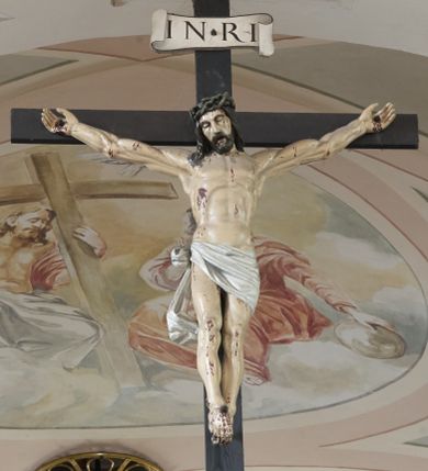 Zdjęcie nr 1: Krucyfiks to przedstawienie ukrzyżowanego Jezusa. Martwe ciało Zbawiciela o wyraźnie zaznaczonej muskulaturze jest rozciągnięte i przybite trzema gwoździami do prostego krzyża. Widać na nim ślady męki: strużki krwi płynące spod grubej korony cierniowej, przebity bok oraz krwawiące rany. Głowa jest opuszczona na prawe ramię, oczy zamknięte, usta rozchylone. Ręce ma rozpostarte, stopy założone na siebie, przybite jednym gwoździem. Biodra otacza białe perizonium opadające na lewym udzie, związane z prawej strony i opadające miękką fałdą w dół. Na górnym ramieniu krzyża widnieje rozwinięty titulus z majuskułowymi literami „INRI” (Iesus Nazarenus Rex Iudaeorum). 