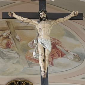 Zdjęcie nr 1: Krucyfiks to przedstawienie ukrzyżowanego Jezusa. Martwe ciało Zbawiciela o wyraźnie zaznaczonej muskulaturze jest rozciągnięte i przybite trzema gwoździami do prostego krzyża. Widać na nim ślady męki: strużki krwi płynące spod grubej korony cierniowej, przebity bok oraz krwawiące rany. Głowa jest opuszczona na prawe ramię, oczy zamknięte, usta rozchylone. Ręce ma rozpostarte, stopy założone na siebie, przybite jednym gwoździem. Biodra otacza białe perizonium opadające na lewym udzie, związane z prawej strony i opadające miękką fałdą w dół. Na górnym ramieniu krzyża widnieje rozwinięty titulus z majuskułowymi literami „INRI” (Iesus Nazarenus Rex Iudaeorum). 