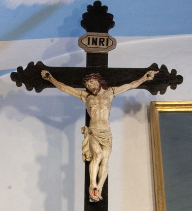 Zdjęcie nr 1: Krucyfiks to przedstawienie ukrzyżowanego Jezusa. Martwe ciało Zbawiciela o wyraźnie zaznaczonej budowie jest rozciągnięte i przybite trzema gwoździami do krzyża, którego ramiona są zakończone wielolistnie. Na ciele widać ślady męki: strużki krwi płynące spod korony cierniowej i ran w dłoniach i stopach oraz przebity bok.  Głowa jest opuszczona na prawe ramię, oczy zamknięte, usta rozchylone. Ręce, których palce są podwinięte, rozpostarte, stopy założone na siebie, przybite jednym gwoździem. Biodra otacza kremowe perizonium opadające na lewym udzie, związane na prawym i opadające miękką, zawijającą się fałdą w dół do wysokości kolan. Na górnym ramieniu krzyża widnieje titulus z majuskułowymi literami „INRI” (Iesus Nazarenus Rex Iudaeorum).