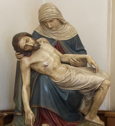 Zdjęcie nr 1: Rzeźba Matki Boskiej trzymającej na kolanach ciało zmarłego Jezusa. Maria siedzi z prawą nogą wystawioną do przodu, opartą na ziemi, lewą stopę ma cofniętą. Prawą rękę trzyma pod głową Jezusa, lewą podtrzymuje Jego lewe przedramię, głowę ma pochyloną do przodu, wzrok kieruje na Syna. Jest ubrana w czerwoną suknię i niebieski, lamowany złotem płaszcz, na głowie ma biały welon. Bezwładne ciało Jezusa opiera się na kolanach Matki, głowa opada na prawy bok, prawa ręka zwisa w kierunki ziemi, lewa, podtrzymywana, ułożona jest równolegle do ciała. Nogi są zgięte w kolanach, oparte na ziemi, na kończynach i w boku widać ślady ran, wokół bioder i ud ma owinięte białe perizonium.