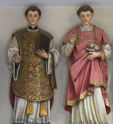 Zdjęcie nr 1: Figura pełna na cokole przedstawiająca św. Szczepana. Święty stoi frontalnie, kierując wzrok w dół. Ma trójkątną twarz, małe oczy, wąski nos i niewielkie usta oraz ciemne, krótki włosy. Prawą rękę, w której brakuje atrybutu (być może gałązki palmowej) unosi przed siebie, lewą przytrzymuje fałdy szaty, w której niesie niewielki stos kamieni. Ubrany jest w białą rokietę zdobioną przy dolnej krawędzi szerokim pasem koronki oraz czerwoną dalmatykę dekorowaną pod szyją i lamowaną złotą taśmą zdobioną przy dolnej krawędzi drogimi kamieniami. Przez lewe przedramię ma przewieszony manipularz. 
Na cokole napis „S[anctus] [Step]han[us]”.