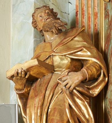 Zdjęcie nr 1: Figura ukazuje mężczyznę w całej postaci, w kontrapoście, ubranego w długą suknię przewiązaną w pasie i płaszcz spięty na piersiach, z otwartą księgą na prawej dłoni, z lewą przytrzymującą na biodrach płaszcz. Głowa zwrócona w lewo, ku górze, okolona długą, falującą brodą. 