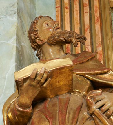 Zdjęcie nr 7: Figura ukazuje mężczyznę w całej postaci, w kontrapoście, ubranego w długą suknię przewiązaną w pasie i płaszcz spięty na piersiach, z otwartą księgą na prawej dłoni, z lewą przytrzymującą na biodrach płaszcz. Głowa zwrócona w lewo, ku górze, okolona długą, falującą brodą. 