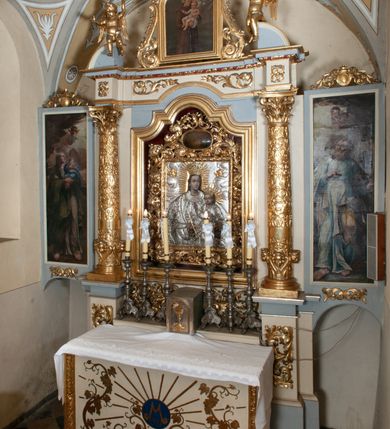 Zdjęcie nr 59: Ołtarz przyścienny, architektoniczny, jednokondygnacyjny ze zwieńczeniem, trójosiowy z bramkami. Mensa murowana prostopadłościenna, antepedium drewniane w złoconej ramie dekorowanej akantem. W jego polu medalion z monogramem maryjnym w glorii promienistej, otoczony przestylizowaną winoroślą. Tabernakulum w osi, metalowe, prostopadłościenne, jednodrzwiczkowe, z przedstawieniem kielicha z hostią. Retabulum ołtarzowe drewniane na cokole. Pole ołtarza z prostokątną, profilowaną ramą zamkniętą łukiem wklęsło-wypukłym z uskokiem. Mieści obraz Matki Boskiej Szkaplerznej, w bogatej akantowo-kwiatowej ramie z umieszczonym u góry, w osi owalnym obrazem Boga Ojca; zasuwany obrazami św. Józefa oraz św. Izydora Oracza. Pole jest ujęte ustawionymi na wysokich cokołach dekorowanych akantem, kolumnami o trzonach wypełnionych ornamentem roślinno-kwiatowym o układzie kandelabrowym oraz u podstawy z pełnoplastycznymi, uskrzydlonymi główkami aniołów; kapitele kompozytowe. Kolumny dźwigają niepełne, wyniesione pośrodku belkowanie, z odcinkami półkolistego przyczółka, na którym siedzą pełnoplastyczne, złocone figurki puttów. Fryz dekorowany akantem. W zwieńczeniu obraz św. Antoniego Padewskiego, prostokątny, zamknięty półkoliście, ujęty akantowymi wolutami. W bocznych osiach, ponad bramkami, prostokątne obrazy z lewej: św. Anny, z prawej: św. Joachima, dekorowane złoconym, ażurowym zwieńczeniem z kartuszy ujętych akantem. Ołtarz utrzymany w odcieniach beżu i szarości ze złoceniami w partiach ornamentów oraz w załamaniach profili architektonicznych.