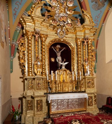 Zdjęcie nr 62: Drewniany, przyścienny ołtarz o formach architektonicznych, jednoosiowy, jednokondygnacyjny ze zwieńczeniem. Mensa murowana w kształcie prostopadłościanu zwieńczonego profilowanym gzymsem; antepedium prostokątne, w drewnianej, płaskorzeźbionej w motywy akantowe, złoconej ramie, dwustronne. Awers tłoczony, srebrzony, wykonany w technice papier mâché; rewers z malowanym na desce wyobrażeniem Veraicon na tle narzędzi męki. Tabernakulum na osi, złożone z mosiężnej, złoconej, sześciobocznej skrzynki w rozbudowanej, drewnianej obudowie o formach architektonicznych. Na nim tron wystawienia w kształcie niszy dekorowanej kratką, nakrytej kopułką z latarnią i przysłoniętej złapanymi po bokach kotarami. Na latarni rzeźbione przedstawienie pelikana karmiącego pisklęta własną krwią. Skrzynkę z tronem ulokowano na tle parawanu ustawionego na podwójnym cokole, ażurowego w górnej części, wypełnionego dekoracją roślinną, złączonego z kopułka spływami. 
Retabulum ołtarzowe drewniane wsparte na wysokim, dwudzielnym, murowanym cokole. Cokół w obudowie drewnianej, jego kondygnacje wydzielone wydatnymi, silnie występującymi przed lico gzymsami; mocno wysunięte ku przodowi części przednie. Ich lica z prostokątnymi płycinami mieszczącymi suchy akant; w części środkowej drugiej kondygnacji cokołu, dodatkowo pełnoplastyczne główki puttów. Na cokole ustawione zdwojone, złocone kolumny o skręconych trzonach i kompozytowych kapitelach. Pomiędzy nimi płyciny wypełnione dekoracją w formie podwieszonego wieńca, u jego szczytu putta pełniące rolę atlantów. Na tle płycin ustawione drewniane, złocone figury z lewej św. Zygmunta i z prawej św. Marii Magdaleny – patronów kościoła. Pole ołtarza z prostokątną niszą zamkniętą łukiem wyniesionym; ujętą płaskorzeźbioną, złoconą ramą zdobioną suchym akantem. W niszy pełnoplastyczny, polichromowany i złocony krucyfiks na aksamitnym tle, flankowany przez płaskorzeźbione wyobrażenia słońca i księżyca. Nisza zasuwana obrazem z Przemienieniem na górze Tabor, w drewnianej, złoconej sukience. Kolumny dźwigają pełne, wyłamane na osiach podpór belkowanie, z wydatnym, silnie profilowanym gzymsem; na jego osi złocony monogram „IHS” w otoku ze srebrzonych obłoków na tle promienistej glorii. Całość wieńczą fragmenty przerwanego przyczółka o formach wklęsło-wypukłych, z ażurową dekoracją z suchego akantu. Pomiędzy ramionami przyczółka złocona gloria promienista, w centrum której płaskorzeźbiona postać Boga Ojca siedzącego na srebrzonym obłoku ze srebrzoną gołębicą Ducha Świętego u stóp. Na ramionach glorii uskrzydlone główki puttów, niektóre na tle obłoków. Po bokach, na gzymsie siedzące postaci nagich, adorujących puttów. Ołtarz ujęty ażurowymi, złoconymi uszami z suchego akantu, w który wplecione są groteskowe putta trzymające pochodnie. Większość powierzchni ołtarza marmoryzowana, w kolorze jasnej, oliwkowej zieleni, z ciemniejszymi brązowymi elementami wypełniającymi płyciny. Złocenia w partiach ornamentów, kolumn oraz załamaniach profili elementów architektonicznych; srebrzone obłoki, części szat postaci i pelikan. 