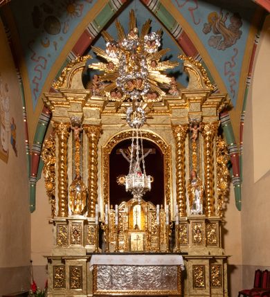 Zdjęcie nr 1: Drewniany, przyścienny ołtarz o formach architektonicznych, jednoosiowy, jednokondygnacyjny ze zwieńczeniem. Mensa murowana w kształcie prostopadłościanu zwieńczonego profilowanym gzymsem; antepedium prostokątne, w drewnianej, płaskorzeźbionej w motywy akantowe, złoconej ramie, dwustronne. Awers tłoczony, srebrzony, wykonany w technice papier mâché; rewers z malowanym na desce wyobrażeniem Veraicon na tle narzędzi męki. Tabernakulum na osi, złożone z mosiężnej, złoconej, sześciobocznej skrzynki w rozbudowanej, drewnianej obudowie o formach architektonicznych. Na nim tron wystawienia w kształcie niszy dekorowanej kratką, nakrytej kopułką z latarnią i przysłoniętej złapanymi po bokach kotarami. Na latarni rzeźbione przedstawienie pelikana karmiącego pisklęta własną krwią. Skrzynkę z tronem ulokowano na tle parawanu ustawionego na podwójnym cokole, ażurowego w górnej części, wypełnionego dekoracją roślinną, złączonego z kopułka spływami. 
Retabulum ołtarzowe drewniane wsparte na wysokim, dwudzielnym, murowanym cokole. Cokół w obudowie drewnianej, jego kondygnacje wydzielone wydatnymi, silnie występującymi przed lico gzymsami; mocno wysunięte ku przodowi części przednie. Ich lica z prostokątnymi płycinami mieszczącymi suchy akant; w części środkowej drugiej kondygnacji cokołu, dodatkowo pełnoplastyczne główki puttów. Na cokole ustawione zdwojone, złocone kolumny o skręconych trzonach i kompozytowych kapitelach. Pomiędzy nimi płyciny wypełnione dekoracją w formie podwieszonego wieńca, u jego szczytu putta pełniące rolę atlantów. Na tle płycin ustawione drewniane, złocone figury z lewej św. Zygmunta i z prawej św. Marii Magdaleny – patronów kościoła. Pole ołtarza z prostokątną niszą zamkniętą łukiem wyniesionym; ujętą płaskorzeźbioną, złoconą ramą zdobioną suchym akantem. W niszy pełnoplastyczny, polichromowany i złocony krucyfiks na aksamitnym tle, flankowany przez płaskorzeźbione wyobrażenia słońca i księżyca. Nisza zasuwana obrazem z Przemienieniem na górze Tabor, w drewnianej, złoconej sukience. Kolumny dźwigają pełne, wyłamane na osiach podpór belkowanie, z wydatnym, silnie profilowanym gzymsem; na jego osi złocony monogram „IHS” w otoku ze srebrzonych obłoków na tle promienistej glorii. Całość wieńczą fragmenty przerwanego przyczółka o formach wklęsło-wypukłych, z ażurową dekoracją z suchego akantu. Pomiędzy ramionami przyczółka złocona gloria promienista, w centrum której płaskorzeźbiona postać Boga Ojca siedzącego na srebrzonym obłoku ze srebrzoną gołębicą Ducha Świętego u stóp. Na ramionach glorii uskrzydlone główki puttów, niektóre na tle obłoków. Po bokach, na gzymsie siedzące postaci nagich, adorujących puttów. Ołtarz ujęty ażurowymi, złoconymi uszami z suchego akantu, w który wplecione są groteskowe putta trzymające pochodnie. Większość powierzchni ołtarza marmoryzowana, w kolorze jasnej, oliwkowej zieleni, z ciemniejszymi brązowymi elementami wypełniającymi płyciny. Złocenia w partiach ornamentów, kolumn oraz załamaniach profili elementów architektonicznych; srebrzone obłoki, części szat postaci i pelikan. 