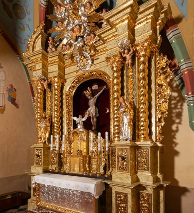 Zdjęcie nr 64: Drewniany, przyścienny ołtarz o formach architektonicznych, jednoosiowy, jednokondygnacyjny ze zwieńczeniem. Mensa murowana w kształcie prostopadłościanu zwieńczonego profilowanym gzymsem; antepedium prostokątne, w drewnianej, płaskorzeźbionej w motywy akantowe, złoconej ramie, dwustronne. Awers tłoczony, srebrzony, wykonany w technice papier mâché; rewers z malowanym na desce wyobrażeniem Veraicon na tle narzędzi męki. Tabernakulum na osi, złożone z mosiężnej, złoconej, sześciobocznej skrzynki w rozbudowanej, drewnianej obudowie o formach architektonicznych. Na nim tron wystawienia w kształcie niszy dekorowanej kratką, nakrytej kopułką z latarnią i przysłoniętej złapanymi po bokach kotarami. Na latarni rzeźbione przedstawienie pelikana karmiącego pisklęta własną krwią. Skrzynkę z tronem ulokowano na tle parawanu ustawionego na podwójnym cokole, ażurowego w górnej części, wypełnionego dekoracją roślinną, złączonego z kopułka spływami. 
Retabulum ołtarzowe drewniane wsparte na wysokim, dwudzielnym, murowanym cokole. Cokół w obudowie drewnianej, jego kondygnacje wydzielone wydatnymi, silnie występującymi przed lico gzymsami; mocno wysunięte ku przodowi części przednie. Ich lica z prostokątnymi płycinami mieszczącymi suchy akant; w części środkowej drugiej kondygnacji cokołu, dodatkowo pełnoplastyczne główki puttów. Na cokole ustawione zdwojone, złocone kolumny o skręconych trzonach i kompozytowych kapitelach. Pomiędzy nimi płyciny wypełnione dekoracją w formie podwieszonego wieńca, u jego szczytu putta pełniące rolę atlantów. Na tle płycin ustawione drewniane, złocone figury z lewej św. Zygmunta i z prawej św. Marii Magdaleny – patronów kościoła. Pole ołtarza z prostokątną niszą zamkniętą łukiem wyniesionym; ujętą płaskorzeźbioną, złoconą ramą zdobioną suchym akantem. W niszy pełnoplastyczny, polichromowany i złocony krucyfiks na aksamitnym tle, flankowany przez płaskorzeźbione wyobrażenia słońca i księżyca. Nisza zasuwana obrazem z Przemienieniem na górze Tabor, w drewnianej, złoconej sukience. Kolumny dźwigają pełne, wyłamane na osiach podpór belkowanie, z wydatnym, silnie profilowanym gzymsem; na jego osi złocony monogram „IHS” w otoku ze srebrzonych obłoków na tle promienistej glorii. Całość wieńczą fragmenty przerwanego przyczółka o formach wklęsło-wypukłych, z ażurową dekoracją z suchego akantu. Pomiędzy ramionami przyczółka złocona gloria promienista, w centrum której płaskorzeźbiona postać Boga Ojca siedzącego na srebrzonym obłoku ze srebrzoną gołębicą Ducha Świętego u stóp. Na ramionach glorii uskrzydlone główki puttów, niektóre na tle obłoków. Po bokach, na gzymsie siedzące postaci nagich, adorujących puttów. Ołtarz ujęty ażurowymi, złoconymi uszami z suchego akantu, w który wplecione są groteskowe putta trzymające pochodnie. Większość powierzchni ołtarza marmoryzowana, w kolorze jasnej, oliwkowej zieleni, z ciemniejszymi brązowymi elementami wypełniającymi płyciny. Złocenia w partiach ornamentów, kolumn oraz załamaniach profili elementów architektonicznych; srebrzone obłoki, części szat postaci i pelikan. 