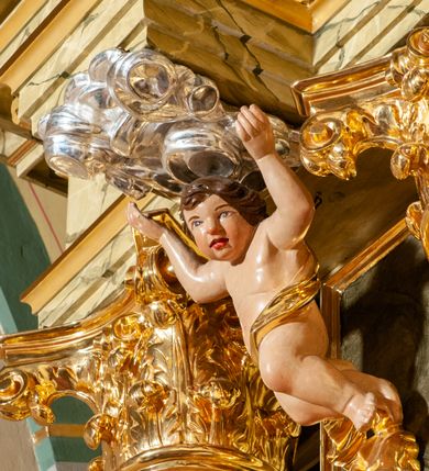 Zdjęcie nr 26: Drewniany, przyścienny ołtarz o formach architektonicznych, jednoosiowy, jednokondygnacyjny ze zwieńczeniem. Mensa murowana w kształcie prostopadłościanu zwieńczonego profilowanym gzymsem; antepedium prostokątne, w drewnianej, płaskorzeźbionej w motywy akantowe, złoconej ramie, dwustronne. Awers tłoczony, srebrzony, wykonany w technice papier mâché; rewers z malowanym na desce wyobrażeniem Veraicon na tle narzędzi męki. Tabernakulum na osi, złożone z mosiężnej, złoconej, sześciobocznej skrzynki w rozbudowanej, drewnianej obudowie o formach architektonicznych. Na nim tron wystawienia w kształcie niszy dekorowanej kratką, nakrytej kopułką z latarnią i przysłoniętej złapanymi po bokach kotarami. Na latarni rzeźbione przedstawienie pelikana karmiącego pisklęta własną krwią. Skrzynkę z tronem ulokowano na tle parawanu ustawionego na podwójnym cokole, ażurowego w górnej części, wypełnionego dekoracją roślinną, złączonego z kopułka spływami. 
Retabulum ołtarzowe drewniane wsparte na wysokim, dwudzielnym, murowanym cokole. Cokół w obudowie drewnianej, jego kondygnacje wydzielone wydatnymi, silnie występującymi przed lico gzymsami; mocno wysunięte ku przodowi części przednie. Ich lica z prostokątnymi płycinami mieszczącymi suchy akant; w części środkowej drugiej kondygnacji cokołu, dodatkowo pełnoplastyczne główki puttów. Na cokole ustawione zdwojone, złocone kolumny o skręconych trzonach i kompozytowych kapitelach. Pomiędzy nimi płyciny wypełnione dekoracją w formie podwieszonego wieńca, u jego szczytu putta pełniące rolę atlantów. Na tle płycin ustawione drewniane, złocone figury z lewej św. Zygmunta i z prawej św. Marii Magdaleny – patronów kościoła. Pole ołtarza z prostokątną niszą zamkniętą łukiem wyniesionym; ujętą płaskorzeźbioną, złoconą ramą zdobioną suchym akantem. W niszy pełnoplastyczny, polichromowany i złocony krucyfiks na aksamitnym tle, flankowany przez płaskorzeźbione wyobrażenia słońca i księżyca. Nisza zasuwana obrazem z Przemienieniem na górze Tabor, w drewnianej, złoconej sukience. Kolumny dźwigają pełne, wyłamane na osiach podpór belkowanie, z wydatnym, silnie profilowanym gzymsem; na jego osi złocony monogram „IHS” w otoku ze srebrzonych obłoków na tle promienistej glorii. Całość wieńczą fragmenty przerwanego przyczółka o formach wklęsło-wypukłych, z ażurową dekoracją z suchego akantu. Pomiędzy ramionami przyczółka złocona gloria promienista, w centrum której płaskorzeźbiona postać Boga Ojca siedzącego na srebrzonym obłoku ze srebrzoną gołębicą Ducha Świętego u stóp. Na ramionach glorii uskrzydlone główki puttów, niektóre na tle obłoków. Po bokach, na gzymsie siedzące postaci nagich, adorujących puttów. Ołtarz ujęty ażurowymi, złoconymi uszami z suchego akantu, w który wplecione są groteskowe putta trzymające pochodnie. Większość powierzchni ołtarza marmoryzowana, w kolorze jasnej, oliwkowej zieleni, z ciemniejszymi brązowymi elementami wypełniającymi płyciny. Złocenia w partiach ornamentów, kolumn oraz załamaniach profili elementów architektonicznych; srebrzone obłoki, części szat postaci i pelikan. 