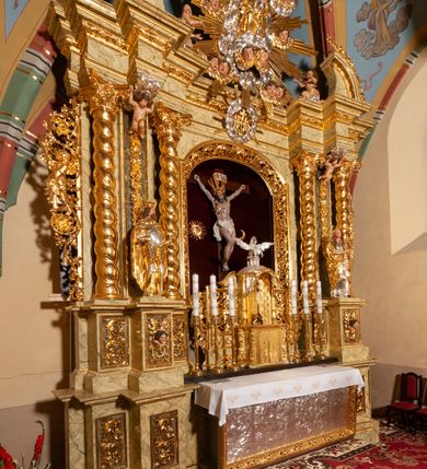 Zdjęcie nr 63: Drewniany, przyścienny ołtarz o formach architektonicznych, jednoosiowy, jednokondygnacyjny ze zwieńczeniem. Mensa murowana w kształcie prostopadłościanu zwieńczonego profilowanym gzymsem; antepedium prostokątne, w drewnianej, płaskorzeźbionej w motywy akantowe, złoconej ramie, dwustronne. Awers tłoczony, srebrzony, wykonany w technice papier mâché; rewers z malowanym na desce wyobrażeniem Veraicon na tle narzędzi męki. Tabernakulum na osi, złożone z mosiężnej, złoconej, sześciobocznej skrzynki w rozbudowanej, drewnianej obudowie o formach architektonicznych. Na nim tron wystawienia w kształcie niszy dekorowanej kratką, nakrytej kopułką z latarnią i przysłoniętej złapanymi po bokach kotarami. Na latarni rzeźbione przedstawienie pelikana karmiącego pisklęta własną krwią. Skrzynkę z tronem ulokowano na tle parawanu ustawionego na podwójnym cokole, ażurowego w górnej części, wypełnionego dekoracją roślinną, złączonego z kopułka spływami. 
Retabulum ołtarzowe drewniane wsparte na wysokim, dwudzielnym, murowanym cokole. Cokół w obudowie drewnianej, jego kondygnacje wydzielone wydatnymi, silnie występującymi przed lico gzymsami; mocno wysunięte ku przodowi części przednie. Ich lica z prostokątnymi płycinami mieszczącymi suchy akant; w części środkowej drugiej kondygnacji cokołu, dodatkowo pełnoplastyczne główki puttów. Na cokole ustawione zdwojone, złocone kolumny o skręconych trzonach i kompozytowych kapitelach. Pomiędzy nimi płyciny wypełnione dekoracją w formie podwieszonego wieńca, u jego szczytu putta pełniące rolę atlantów. Na tle płycin ustawione drewniane, złocone figury z lewej św. Zygmunta i z prawej św. Marii Magdaleny – patronów kościoła. Pole ołtarza z prostokątną niszą zamkniętą łukiem wyniesionym; ujętą płaskorzeźbioną, złoconą ramą zdobioną suchym akantem. W niszy pełnoplastyczny, polichromowany i złocony krucyfiks na aksamitnym tle, flankowany przez płaskorzeźbione wyobrażenia słońca i księżyca. Nisza zasuwana obrazem z Przemienieniem na górze Tabor, w drewnianej, złoconej sukience. Kolumny dźwigają pełne, wyłamane na osiach podpór belkowanie, z wydatnym, silnie profilowanym gzymsem; na jego osi złocony monogram „IHS” w otoku ze srebrzonych obłoków na tle promienistej glorii. Całość wieńczą fragmenty przerwanego przyczółka o formach wklęsło-wypukłych, z ażurową dekoracją z suchego akantu. Pomiędzy ramionami przyczółka złocona gloria promienista, w centrum której płaskorzeźbiona postać Boga Ojca siedzącego na srebrzonym obłoku ze srebrzoną gołębicą Ducha Świętego u stóp. Na ramionach glorii uskrzydlone główki puttów, niektóre na tle obłoków. Po bokach, na gzymsie siedzące postaci nagich, adorujących puttów. Ołtarz ujęty ażurowymi, złoconymi uszami z suchego akantu, w który wplecione są groteskowe putta trzymające pochodnie. Większość powierzchni ołtarza marmoryzowana, w kolorze jasnej, oliwkowej zieleni, z ciemniejszymi brązowymi elementami wypełniającymi płyciny. Złocenia w partiach ornamentów, kolumn oraz załamaniach profili elementów architektonicznych; srebrzone obłoki, części szat postaci i pelikan. 