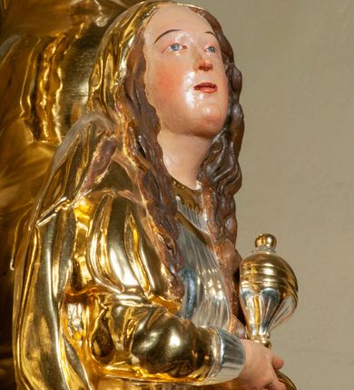 Zdjęcie nr 7: Pełnoplastyczna, drążona od tyłu, drewniana rzeźba stojąca po prawej stronie ołtarza głównego. Maria Magdalena stoi w lekkim kontrapoście, z głową przechyloną na prawo. W prawym ręku trzyma puszkę na wonności, lewą, opuszczoną wzdłuż ciała, podtrzymuje draperie szat. Ma owalną twarz o pełnych policzkach i wysokim czole. Niewielkie oczy i usta, wąskie brwi. Długie, falowane włosy opadają jej na oba ramiona. Jest ubrana w sięgającą ziemi, srebrzoną suknię oraz obfity, długi, złocony płaszcz, którego poła okrywa głowę postaci. U dołu, spod szat, widoczne są czubki jej butów. Włosy, karnacja oraz buty polichromowane; szaty srebrzone i złocone.