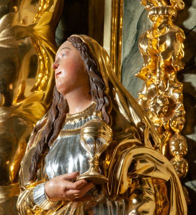 Zdjęcie nr 6: Pełnoplastyczna, drążona od tyłu, drewniana rzeźba stojąca po prawej stronie ołtarza głównego. Maria Magdalena stoi w lekkim kontrapoście, z głową przechyloną na prawo. W prawym ręku trzyma puszkę na wonności, lewą, opuszczoną wzdłuż ciała, podtrzymuje draperie szat. Ma owalną twarz o pełnych policzkach i wysokim czole. Niewielkie oczy i usta, wąskie brwi. Długie, falowane włosy opadają jej na oba ramiona. Jest ubrana w sięgającą ziemi, srebrzoną suknię oraz obfity, długi, złocony płaszcz, którego poła okrywa głowę postaci. U dołu, spod szat, widoczne są czubki jej butów. Włosy, karnacja oraz buty polichromowane; szaty srebrzone i złocone.