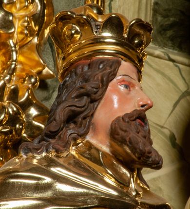 Zdjęcie nr 8: Pełnoplastyczna, wydrążona z tyłu do połowy wysokości, drewniana figura św. Zygmunta, ustawiona po lewej stronie ołtarza głównego. Święty został przedstawiony w pozycji stojącej, z lewą nogą wysuniętą nieco do przodu, ubrany w zbroję płytową, na którą narzucono mu obfity płaszcz spięty pod szyją. W lewej, ugiętej w łokciu i wyciągniętej ku przodowi ręce trzyma jabłko królewskie, prawą wspiera na mieczu przytroczonym do boku. Twarz pociągła, okolona długimi włosami opadającymi na oba ramiona i falującą brodą. Ma pełne, lekko rozchylone usta, prosty nos i przysłonięte grubymi powiekami oczy; na głowie korona. Partie twarzy i włosów polichromowane, szaty i atrybuty władzy królewskiej złocone. Części zbroi (napierśnik i nakolanniki) srebrzone, pozostałe partie złocone.