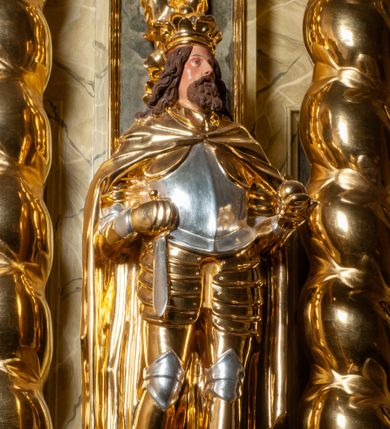 Zdjęcie nr 1: Pełnoplastyczna, wydrążona z tyłu do połowy wysokości, drewniana figura św. Zygmunta, ustawiona po lewej stronie ołtarza głównego. Święty został przedstawiony w pozycji stojącej, z lewą nogą wysuniętą nieco do przodu, ubrany w zbroję płytową, na którą narzucono mu obfity płaszcz spięty pod szyją. W lewej, ugiętej w łokciu i wyciągniętej ku przodowi ręce trzyma jabłko królewskie, prawą wspiera na mieczu przytroczonym do boku. Twarz pociągła, okolona długimi włosami opadającymi na oba ramiona i falującą brodą. Ma pełne, lekko rozchylone usta, prosty nos i przysłonięte grubymi powiekami oczy; na głowie korona. Partie twarzy i włosów polichromowane, szaty i atrybuty władzy królewskiej złocone. Części zbroi (napierśnik i nakolanniki) srebrzone, pozostałe partie złocone.