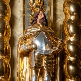 Zdjęcie nr 1: Pełnoplastyczna, wydrążona z tyłu do połowy wysokości, drewniana figura św. Zygmunta, ustawiona po lewej stronie ołtarza głównego. Święty został przedstawiony w pozycji stojącej, z lewą nogą wysuniętą nieco do przodu, ubrany w zbroję płytową, na którą narzucono mu obfity płaszcz spięty pod szyją. W lewej, ugiętej w łokciu i wyciągniętej ku przodowi ręce trzyma jabłko królewskie, prawą wspiera na mieczu przytroczonym do boku. Twarz pociągła, okolona długimi włosami opadającymi na oba ramiona i falującą brodą. Ma pełne, lekko rozchylone usta, prosty nos i przysłonięte grubymi powiekami oczy; na głowie korona. Partie twarzy i włosów polichromowane, szaty i atrybuty władzy królewskiej złocone. Części zbroi (napierśnik i nakolanniki) srebrzone, pozostałe partie złocone.