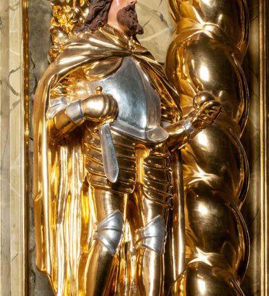 Zdjęcie nr 4: Pełnoplastyczna, wydrążona z tyłu do połowy wysokości, drewniana figura św. Zygmunta, ustawiona po lewej stronie ołtarza głównego. Święty został przedstawiony w pozycji stojącej, z lewą nogą wysuniętą nieco do przodu, ubrany w zbroję płytową, na którą narzucono mu obfity płaszcz spięty pod szyją. W lewej, ugiętej w łokciu i wyciągniętej ku przodowi ręce trzyma jabłko królewskie, prawą wspiera na mieczu przytroczonym do boku. Twarz pociągła, okolona długimi włosami opadającymi na oba ramiona i falującą brodą. Ma pełne, lekko rozchylone usta, prosty nos i przysłonięte grubymi powiekami oczy; na głowie korona. Partie twarzy i włosów polichromowane, szaty i atrybuty władzy królewskiej złocone. Części zbroi (napierśnik i nakolanniki) srebrzone, pozostałe partie złocone.