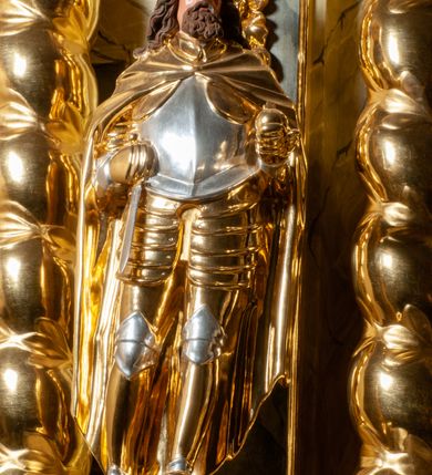 Zdjęcie nr 3: Pełnoplastyczna, wydrążona z tyłu do połowy wysokości, drewniana figura św. Zygmunta, ustawiona po lewej stronie ołtarza głównego. Święty został przedstawiony w pozycji stojącej, z lewą nogą wysuniętą nieco do przodu, ubrany w zbroję płytową, na którą narzucono mu obfity płaszcz spięty pod szyją. W lewej, ugiętej w łokciu i wyciągniętej ku przodowi ręce trzyma jabłko królewskie, prawą wspiera na mieczu przytroczonym do boku. Twarz pociągła, okolona długimi włosami opadającymi na oba ramiona i falującą brodą. Ma pełne, lekko rozchylone usta, prosty nos i przysłonięte grubymi powiekami oczy; na głowie korona. Partie twarzy i włosów polichromowane, szaty i atrybuty władzy królewskiej złocone. Części zbroi (napierśnik i nakolanniki) srebrzone, pozostałe partie złocone.