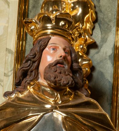 Zdjęcie nr 6: Pełnoplastyczna, wydrążona z tyłu do połowy wysokości, drewniana figura św. Zygmunta, ustawiona po lewej stronie ołtarza głównego. Święty został przedstawiony w pozycji stojącej, z lewą nogą wysuniętą nieco do przodu, ubrany w zbroję płytową, na którą narzucono mu obfity płaszcz spięty pod szyją. W lewej, ugiętej w łokciu i wyciągniętej ku przodowi ręce trzyma jabłko królewskie, prawą wspiera na mieczu przytroczonym do boku. Twarz pociągła, okolona długimi włosami opadającymi na oba ramiona i falującą brodą. Ma pełne, lekko rozchylone usta, prosty nos i przysłonięte grubymi powiekami oczy; na głowie korona. Partie twarzy i włosów polichromowane, szaty i atrybuty władzy królewskiej złocone. Części zbroi (napierśnik i nakolanniki) srebrzone, pozostałe partie złocone.